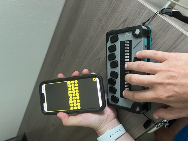 La foto presenta un iPhone con el app BrailleBuzz y una línea Braille.